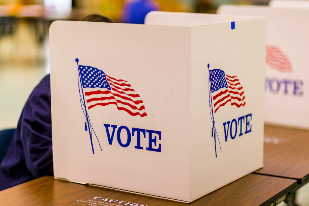 Person at election voting behind booth. Booth reads "vote" with american flag.