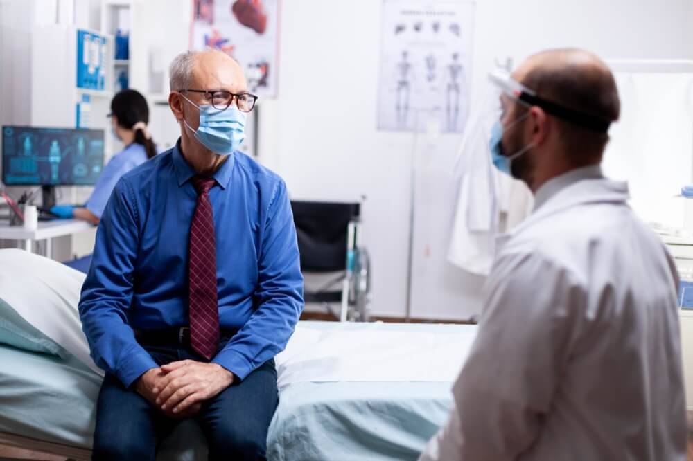 White doctor in PPE talks to white patient wearing surgical mask