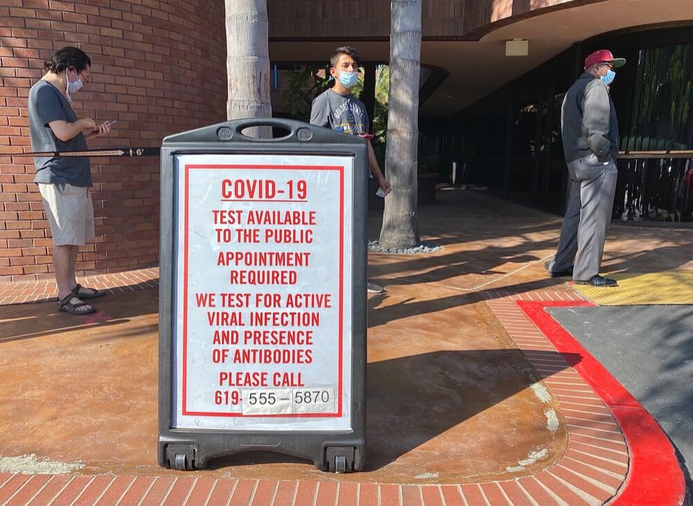 People wait in socially distanced line for COVID-19 test. Sign reads "Covid-19 Test Available To the Public Appointment Required We Test for Active Viral Infection And Presence Of Antibodies"