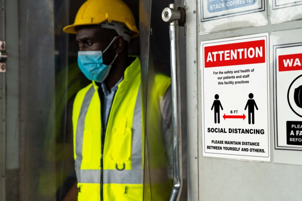 Black construction worker wearing mask and vest. Sign on wall advises about COVID-19 and social distancing.