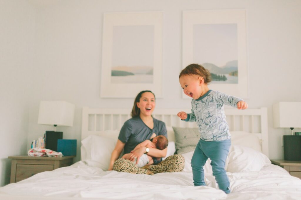 Mother nursing baby while toddler jumps on bed