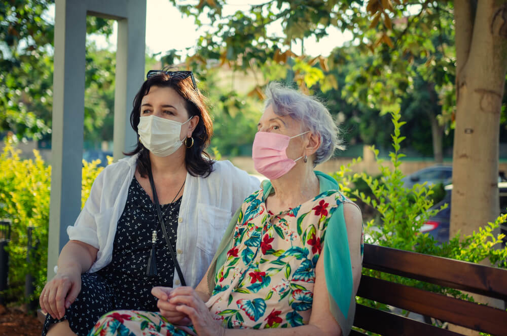 Attentive caregiver or companion and a senior adult woman in protective masks are sitting on a park bench. Summer sunny day.