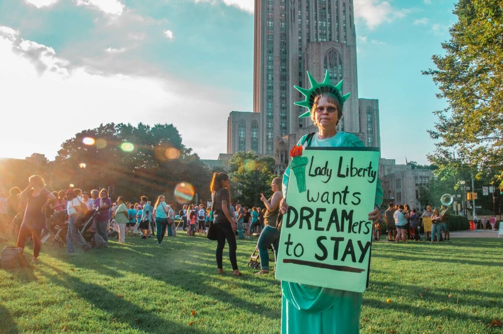 Person dressed as Lady Liberty standing in park with sign; sign reads "Lady Liberty wants DREAMers to stay."