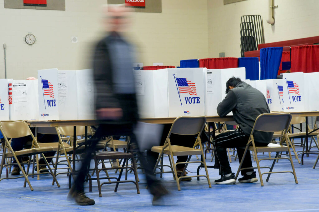 Voter casting ballot in sitdown booth while other voters pass by in foreground