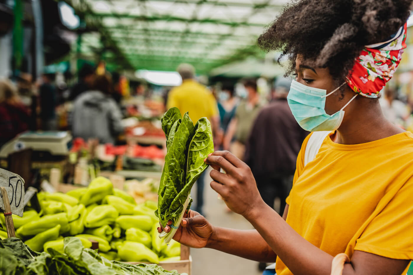 Young African American woman with face mask inspecting kale at farmers market