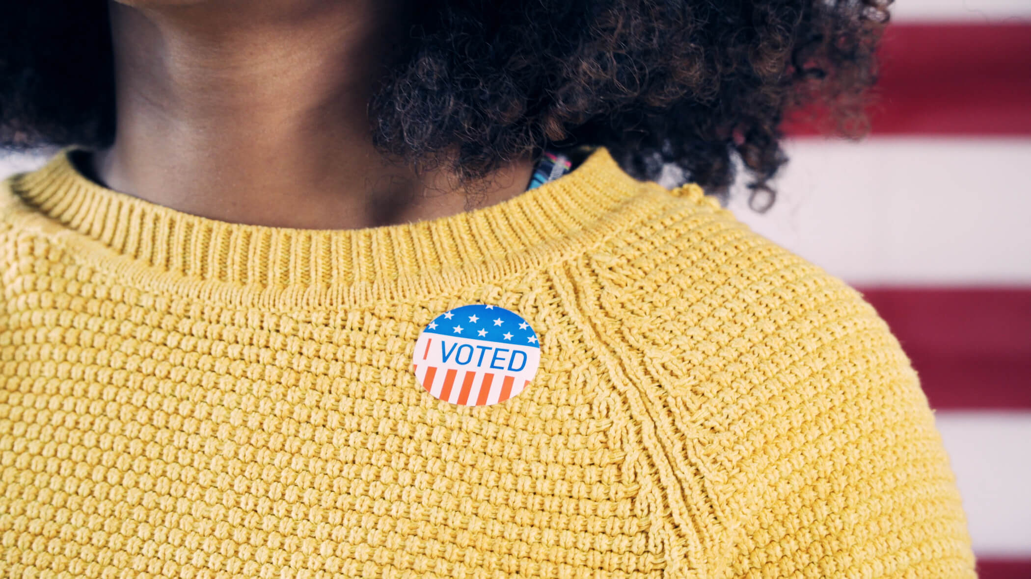 Black person in yellow sweater wearing "I voted" sticker; American flag in background