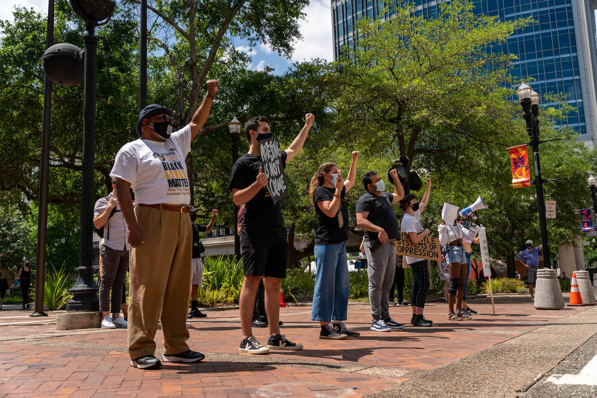 A multiracial group of 6 demonstrators stand with fist raised in front of Jacksonville, Florida city hall. Visible signs read, “Protest is not a crime,” and “Black Lives Matter.”