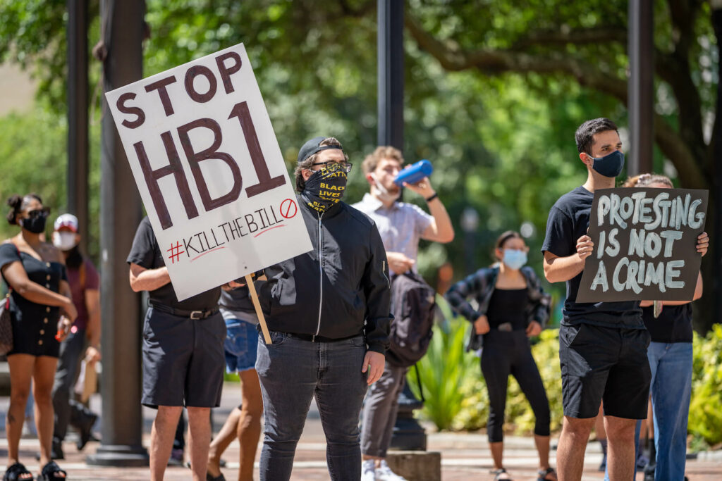 Protests gather to demonstrate against HB1. Two demonstrators in foreground hold signs reading, “Stop HB1 #KillTheBill,” and “Protesting is not a crime.”