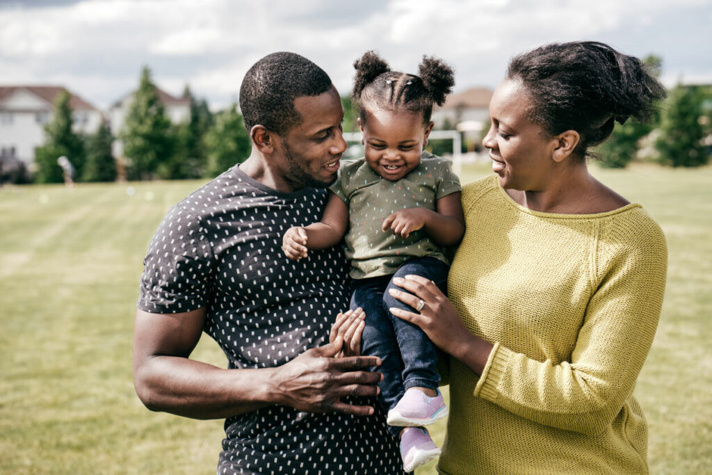 Black mother and Black father playfully hold their toddler daughter in field outdoors