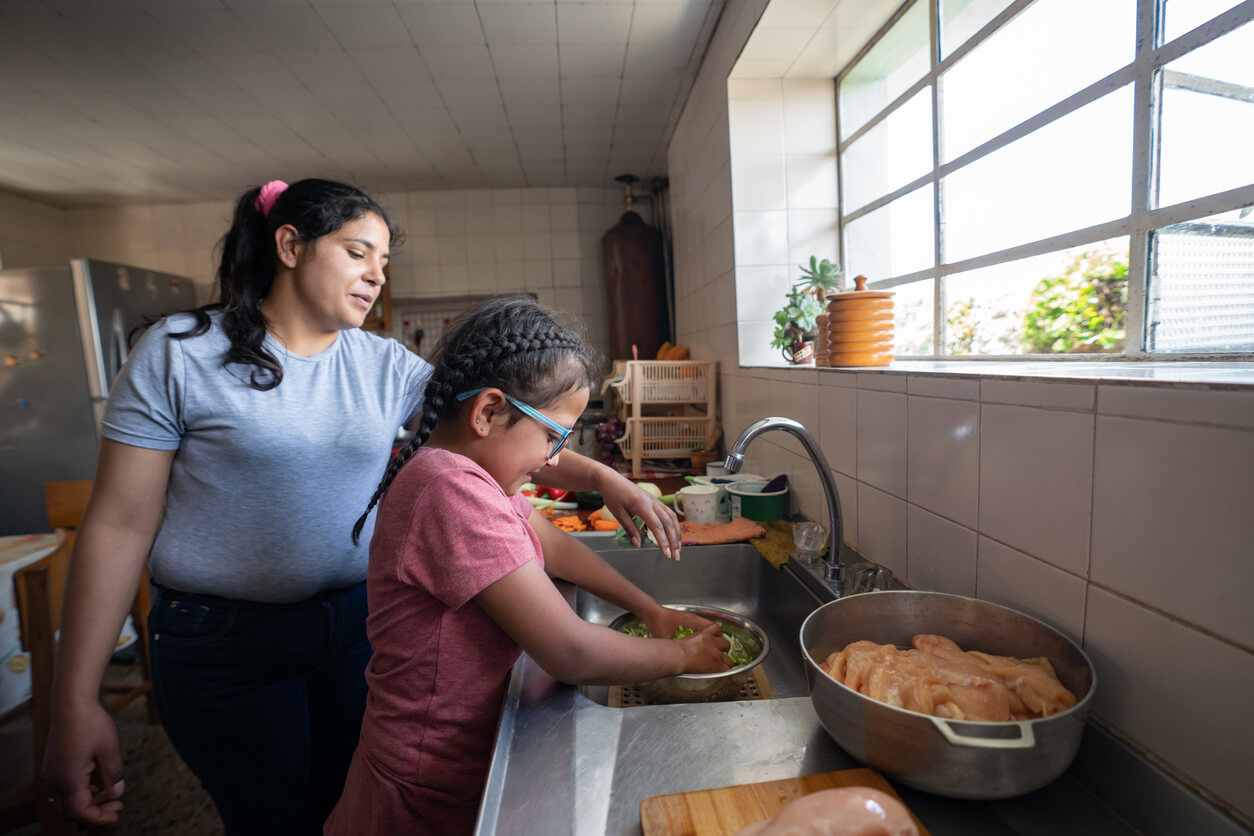 Latina mother teaches daughter how to wash vegetables in kitchen sink