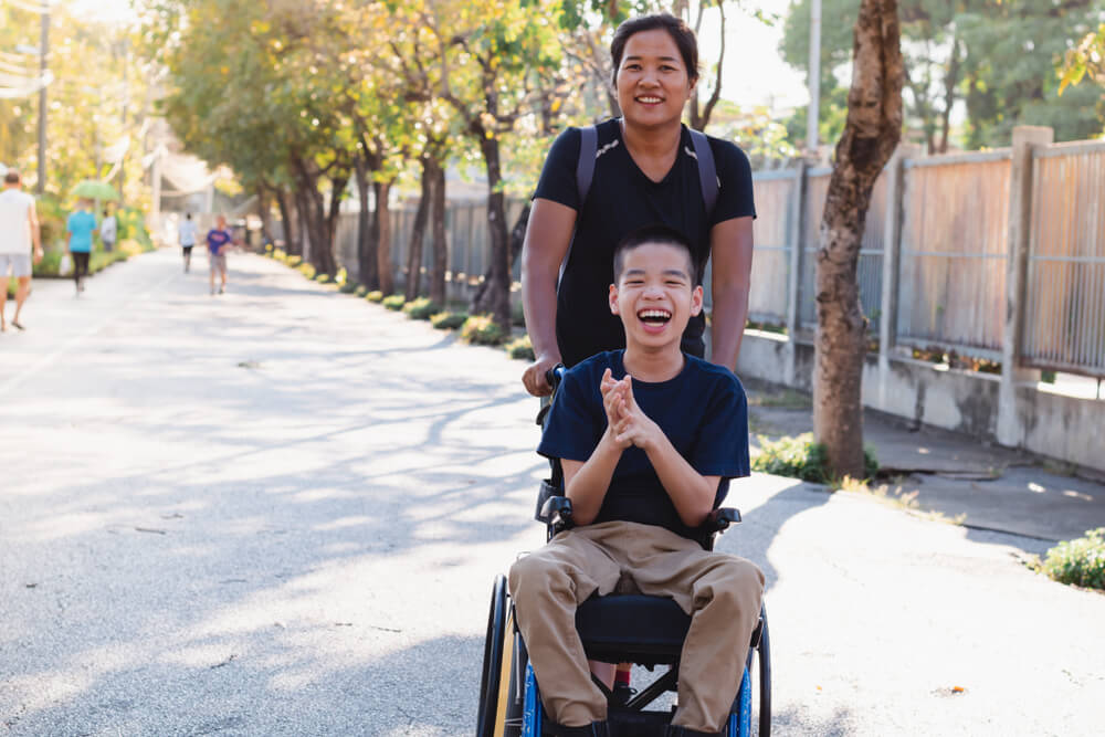 Young Asian child in wheelchair laughs with caretaker or family member at the park