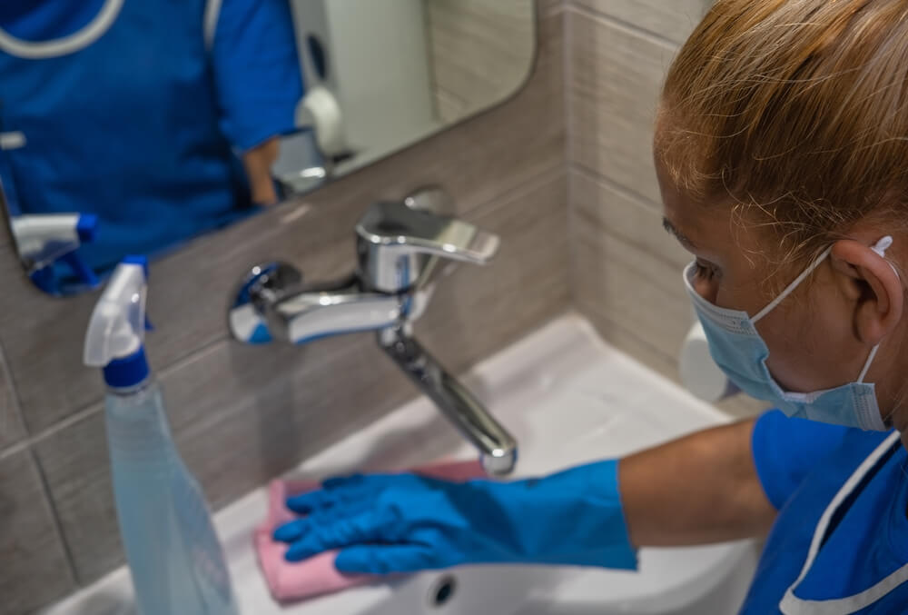 Latina custodian wearing mask cleans bathroom sink or washbasin with towel