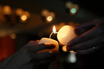 Close up of two people's hands holding white candles with long wicks against blurry background. The hands on the right use their candle to light the candle on the left.